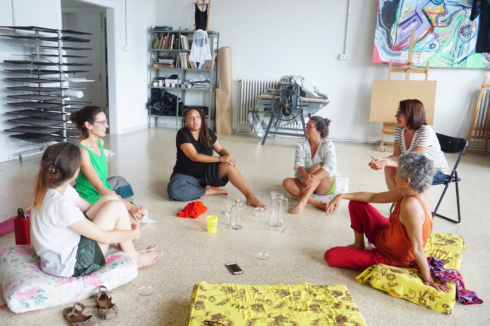 Women seated in a circle on the floor of an art studio, in conversation
